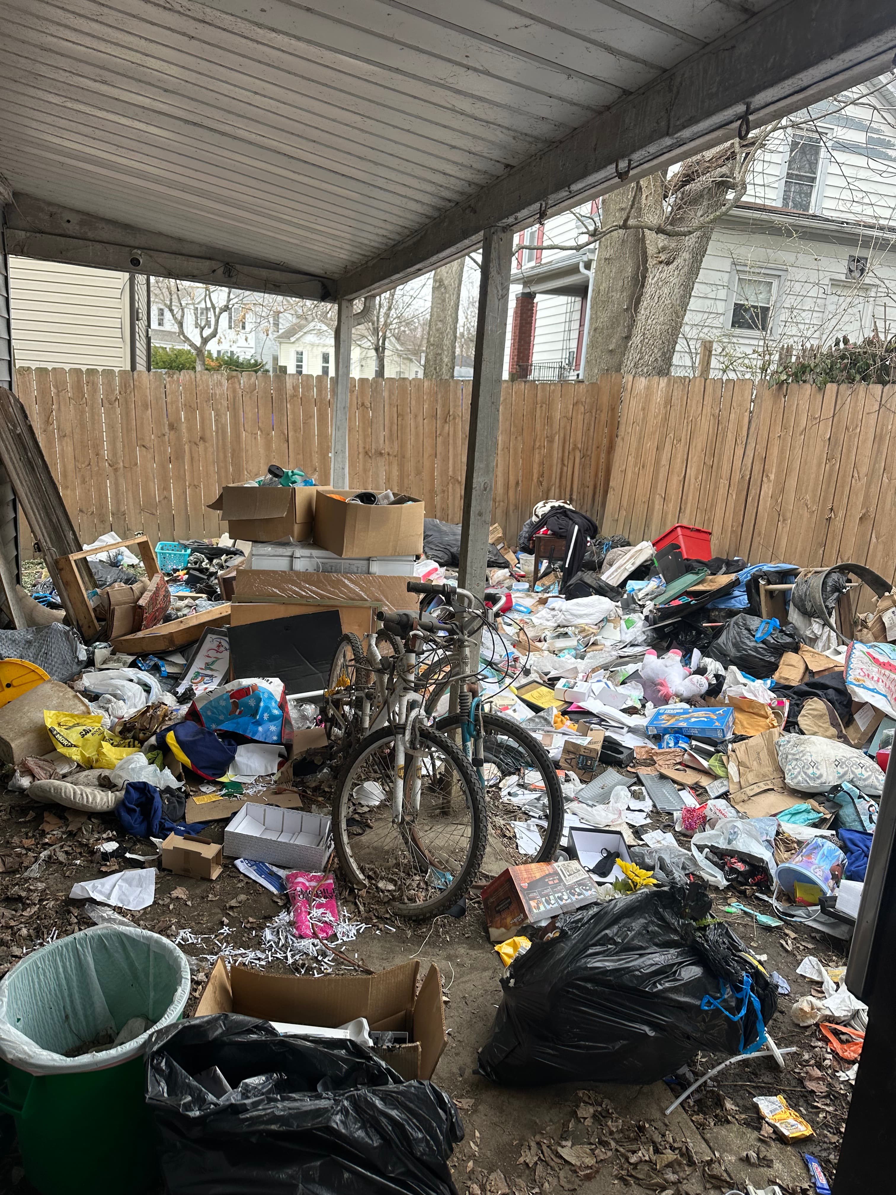 Massive pile of trash, garbage bags, cardboard boxes, and a bicycle against a wooden fence.