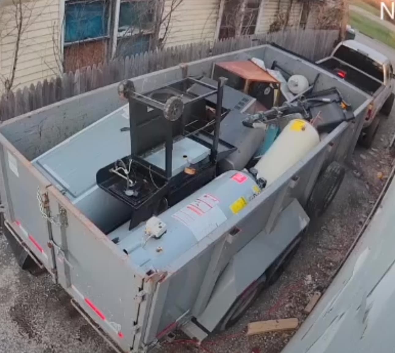Grey utility trailer filled with discarded items including a water heater and a grill.
