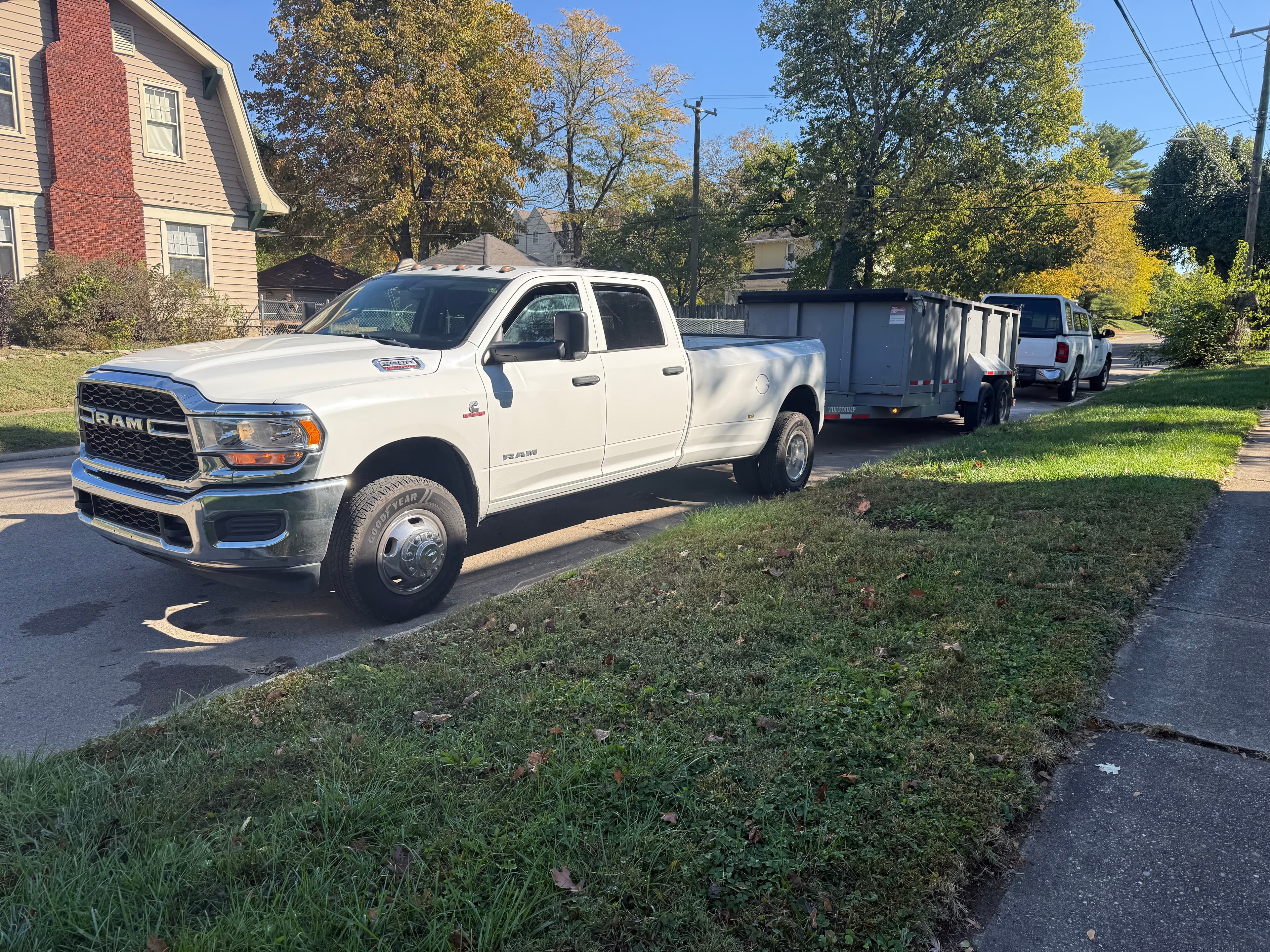 White Ram dually truck towing a grey dump trailer on a sunny residential street.