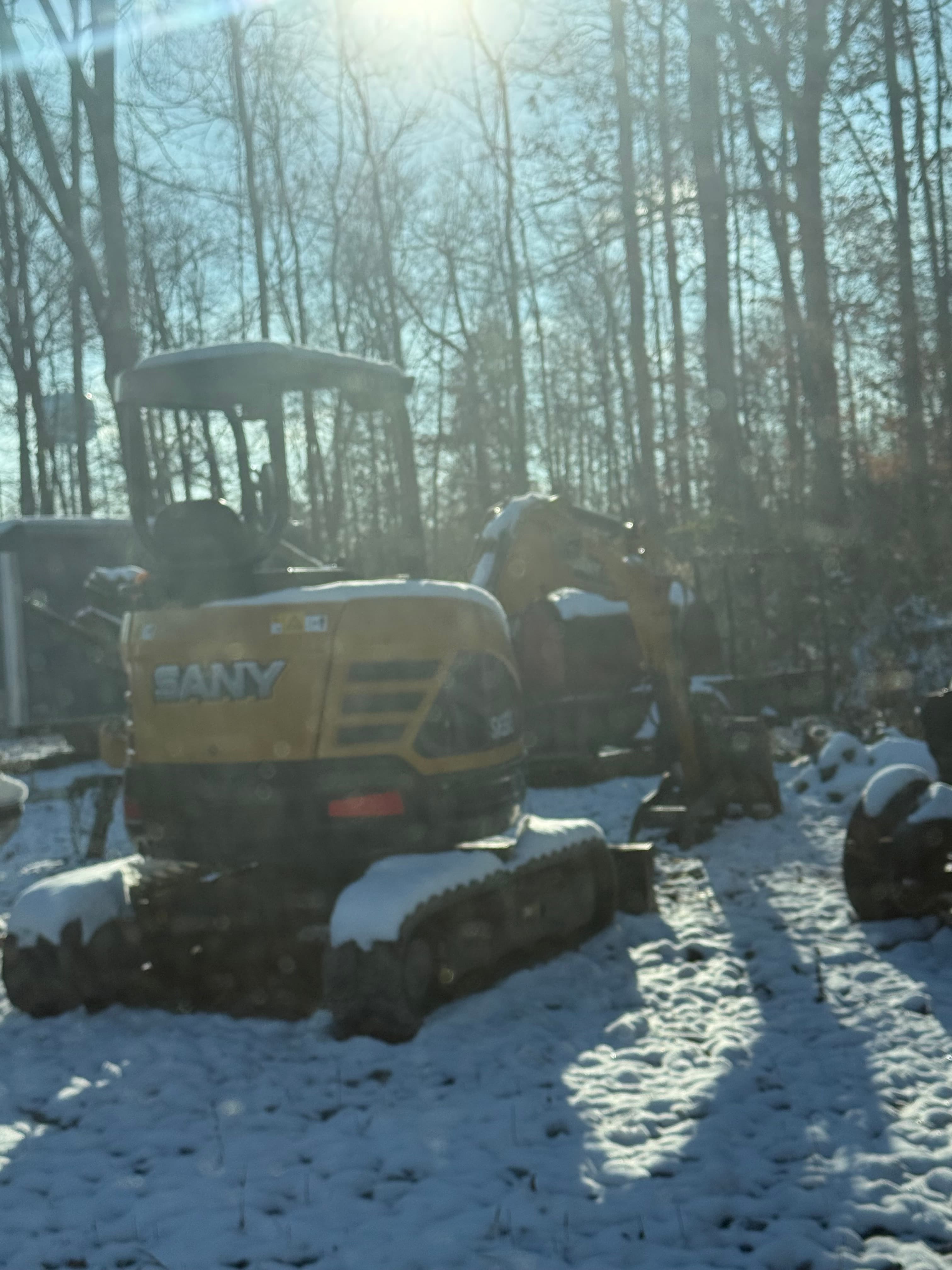 Yellow SANY mini excavator on snowy ground with bare winter trees and bright sunlight behind.