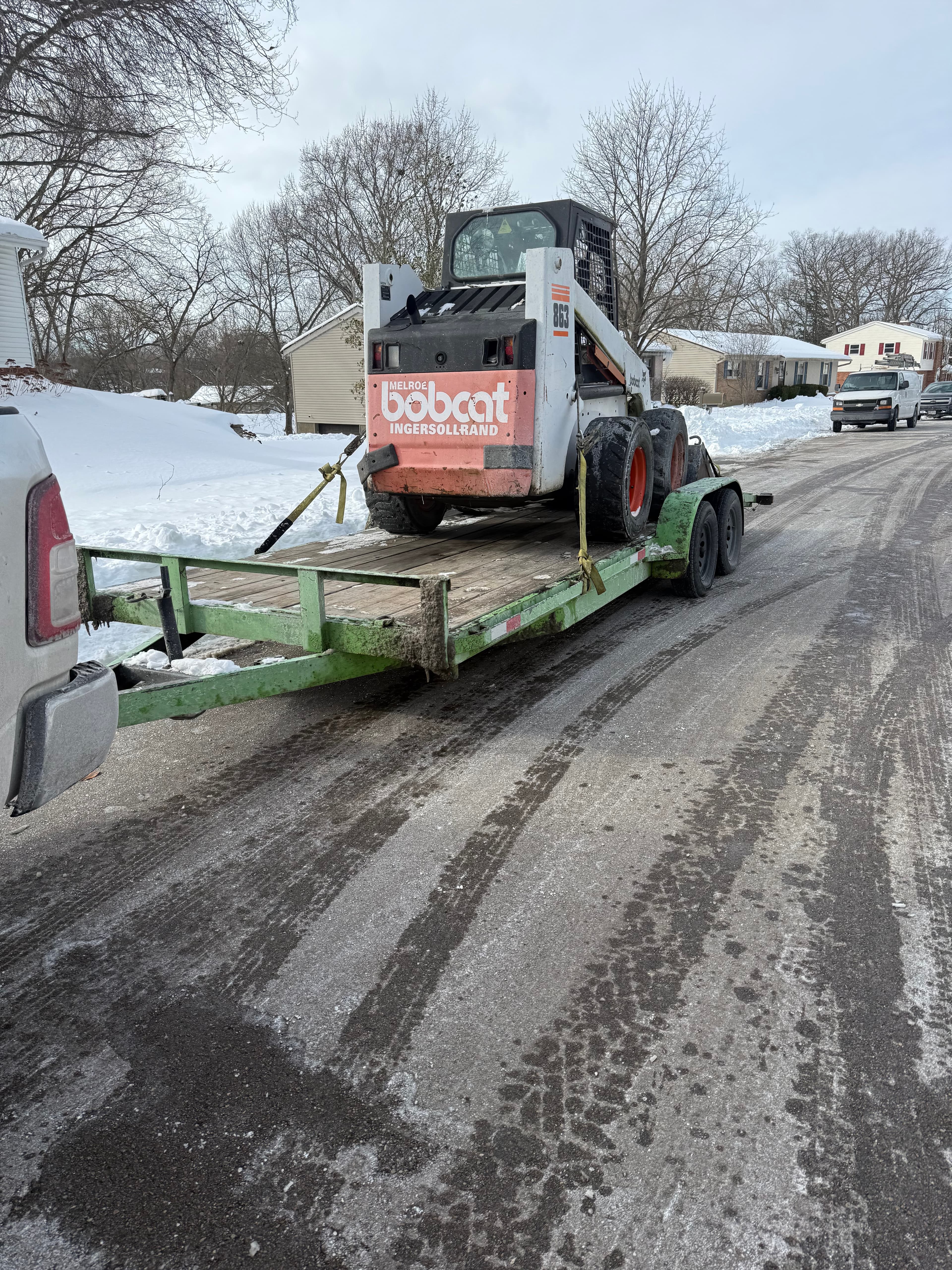 White Bobcat skid-steer loader on a green trailer parked on a snowy residential street.