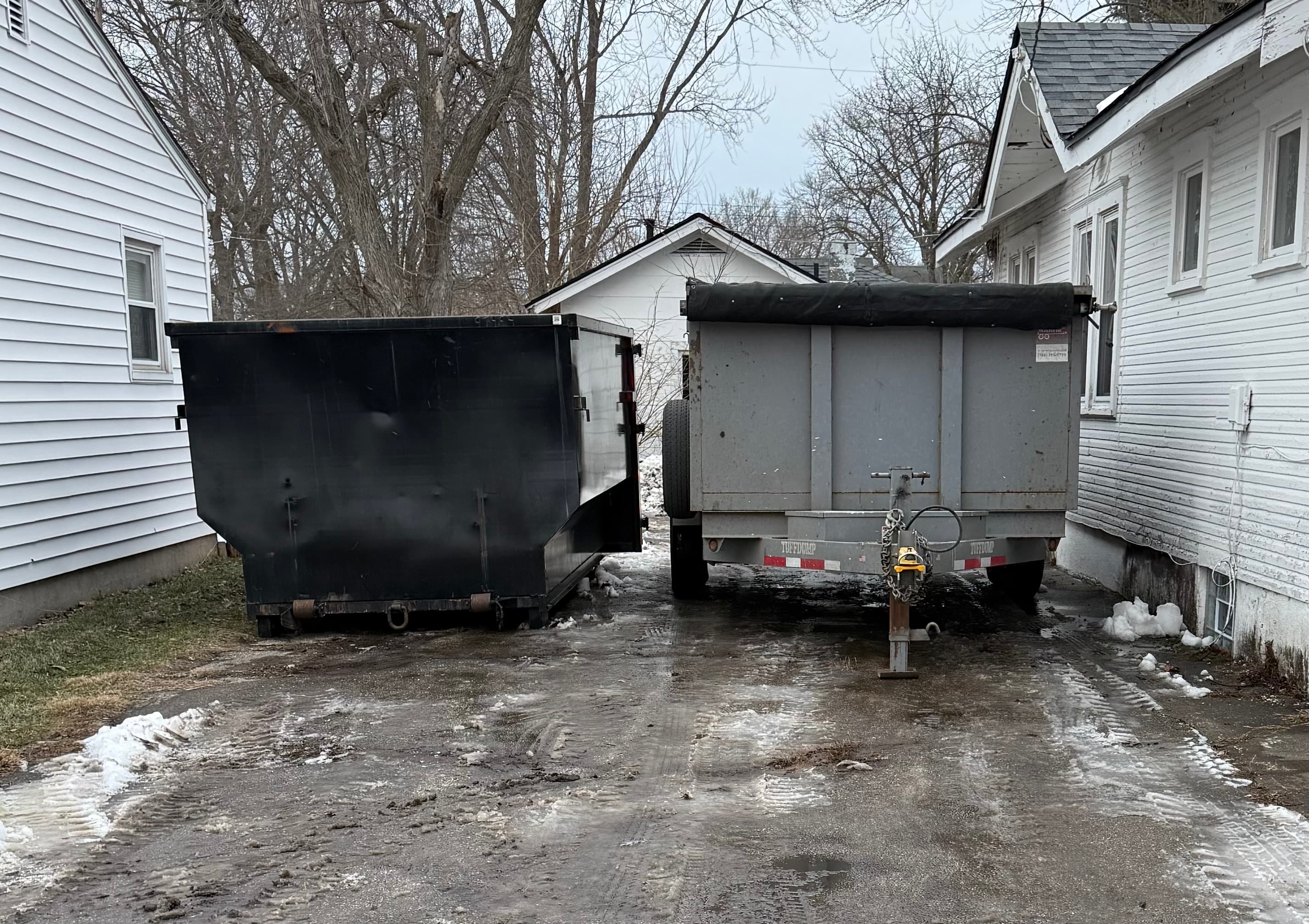 Black dumpster and grey dump trailer parked in a muddy driveway between two white houses.